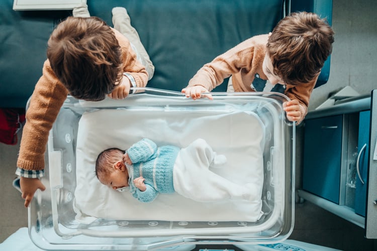Twins Looking At Their New Sibling In A Hospital Bassinet